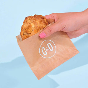 Hand holding a Cereal Milk New York Cookie in Crumbs & Doilies brown paper bag against blue background